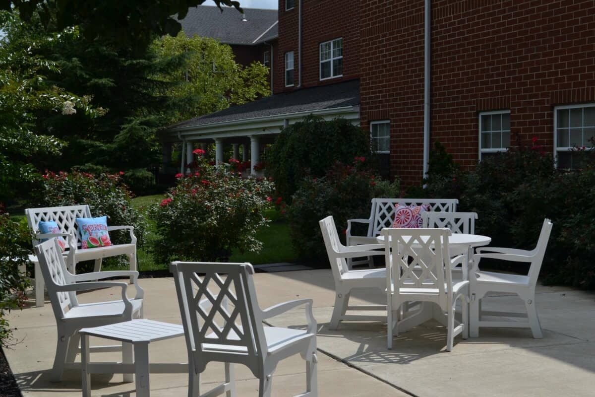 A patio area with white furniture including chairs and a table, set against a brick building and surrounded by greenery.