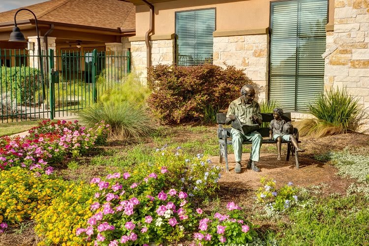 An elderly man sits on a bench surrounded by blooming flowers in a garden.