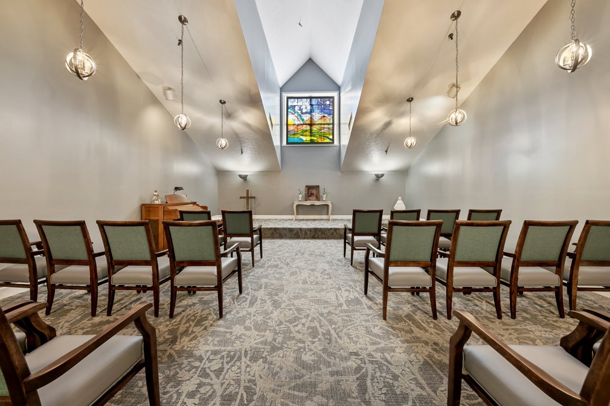 A serene chapel interior featuring rows of chairs, a stained glass window, and an altar at the front.
