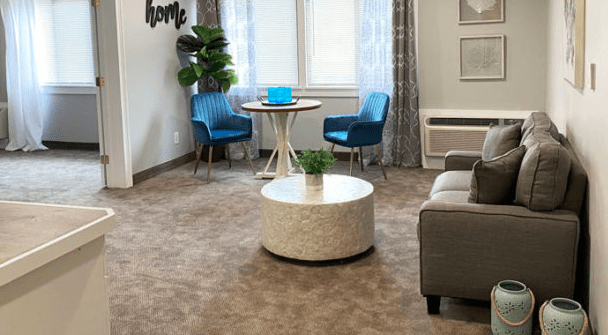 A cozy living room with a small table and two blue chairs by the window, complemented by a round coffee table and a comfortable gray sofa.
