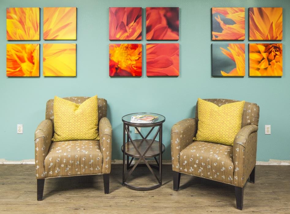 A modern living room setup featuring two patterned armchairs with yellow pillows, a glass-topped side table, and vibrant abstract wall art.