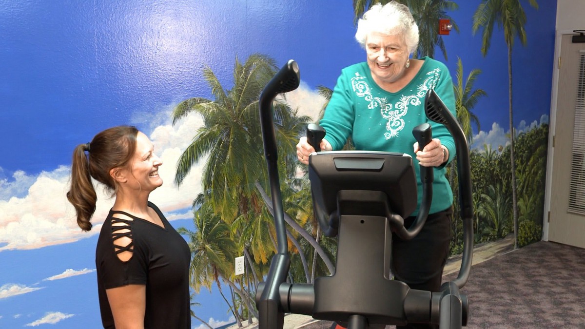 An elderly woman enjoys a workout on exercise equipment while being assisted by a trainer in front of a tropical mural.