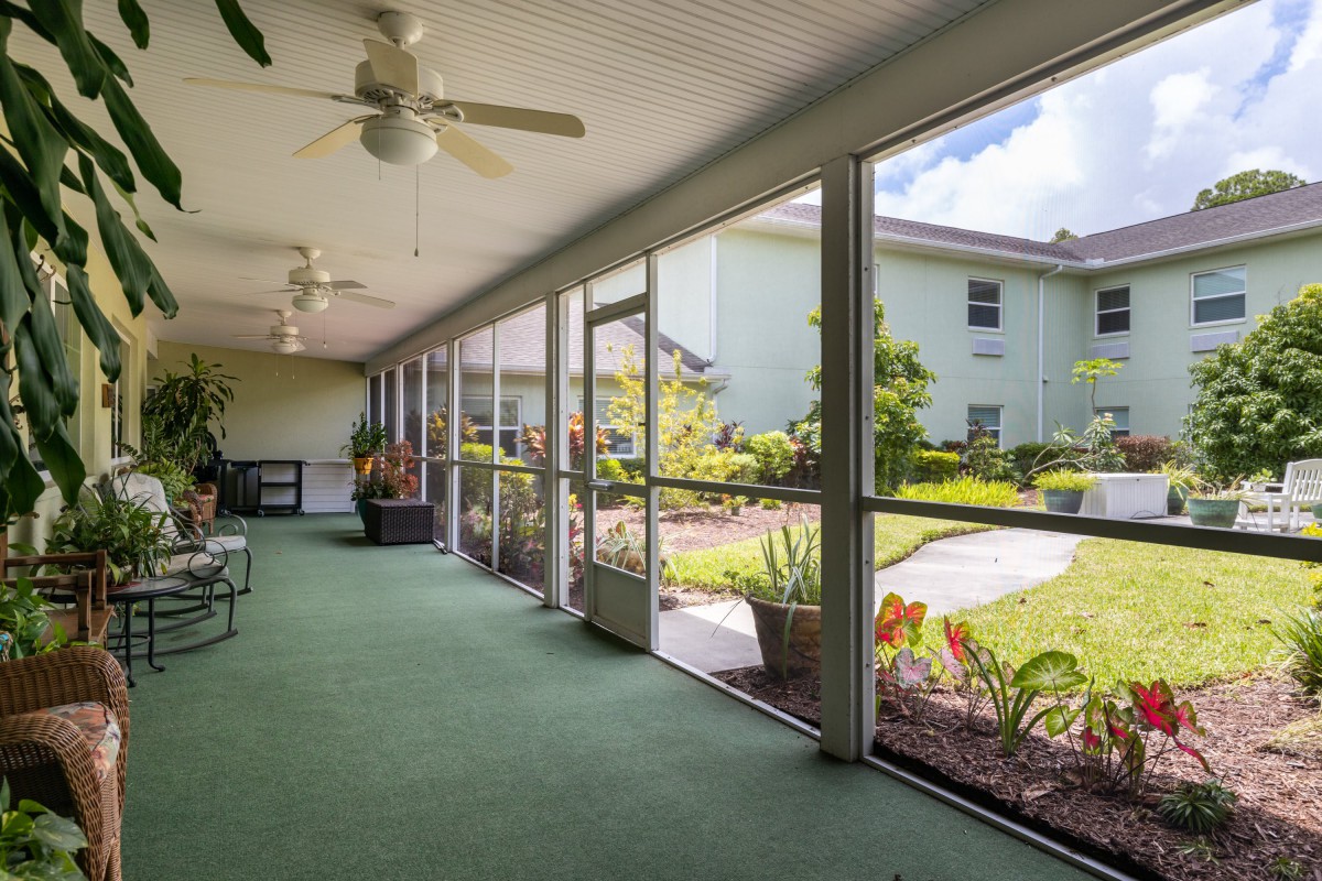 A screened porch with green carpeting, ceiling fans, and outdoor views of a garden space.