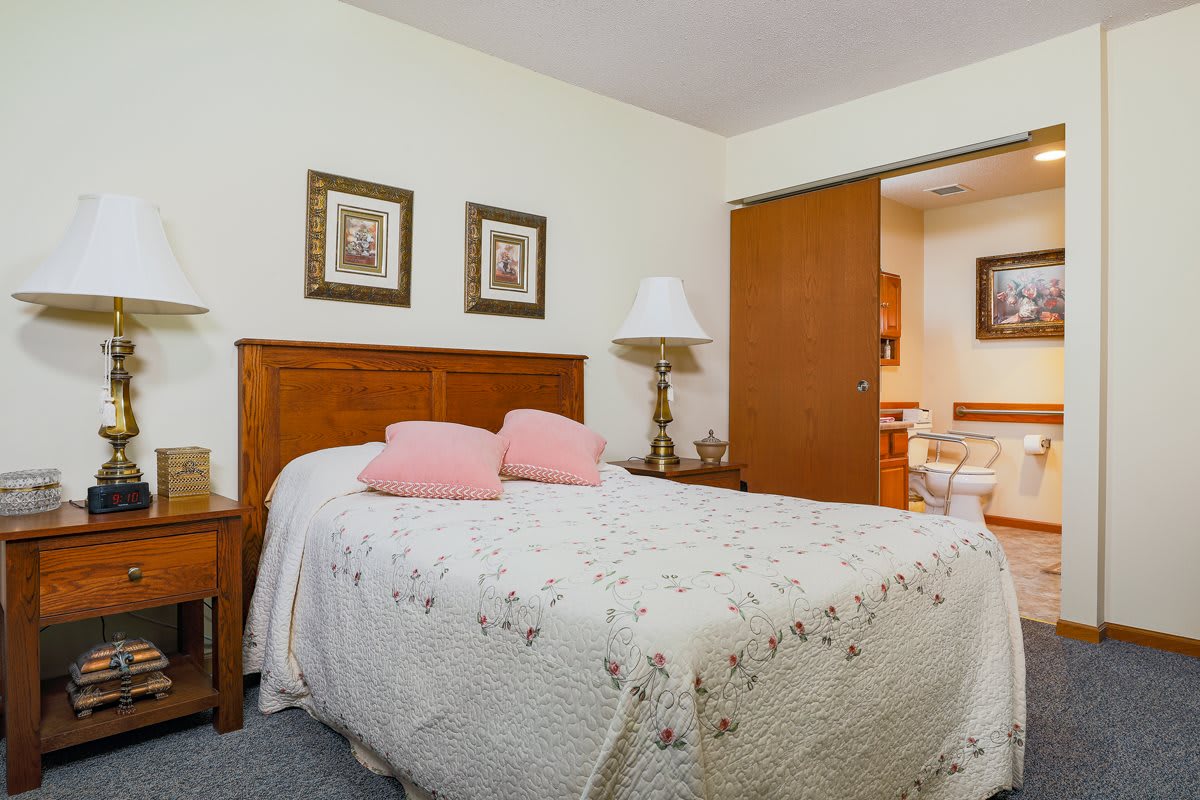 A cozy bedroom featuring a wooden bed with a floral quilt and pink pillows, flanked by nightstands and lamps, with framed artwork on the walls.