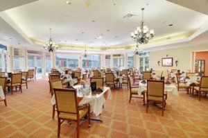 A spacious dining room with elegantly set tables and chairs, featuring chandeliers and large windows.