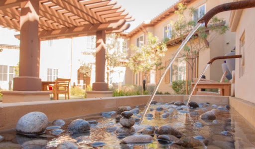A serene outdoor space featuring a beautiful fountain surrounded by pebbles and shaded seating.