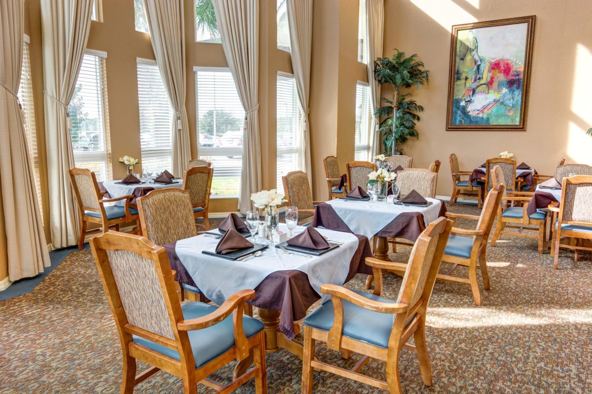 A well-lit dining room with elegantly set tables and wooden chairs, featuring large windows and tasteful decor.