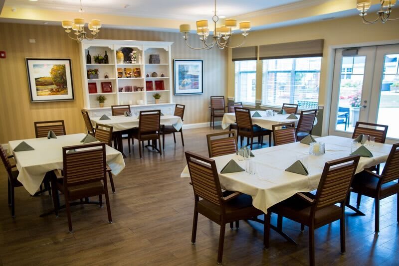A well-lit dining room with neatly arranged tables and chairs, featuring elegant chandeliers and large windows.