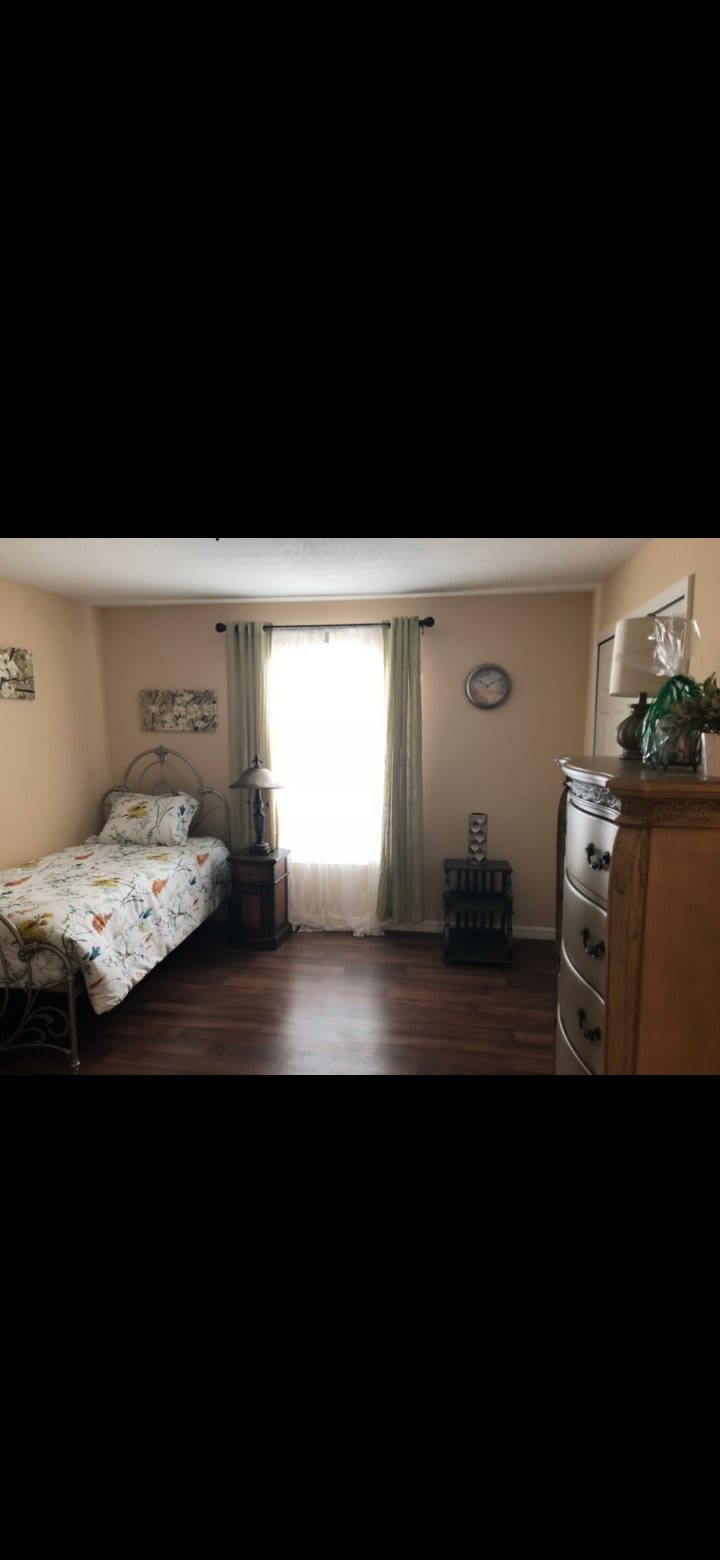 A cozy bedroom with a single bed, wooden dresser, and curtains allowing natural light to fill the room.