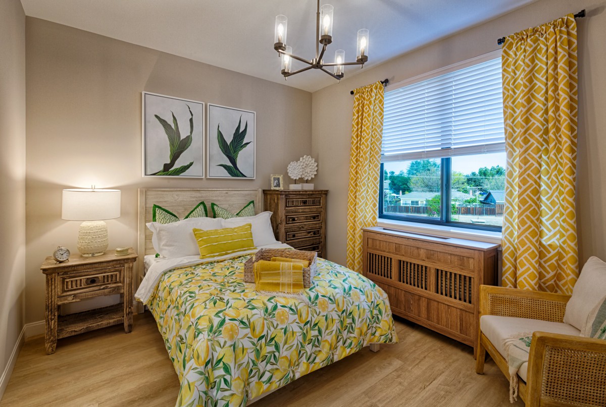 A cozy bedroom with yellow accents and botanical artwork, featuring a neatly made bed and elegant lighting.