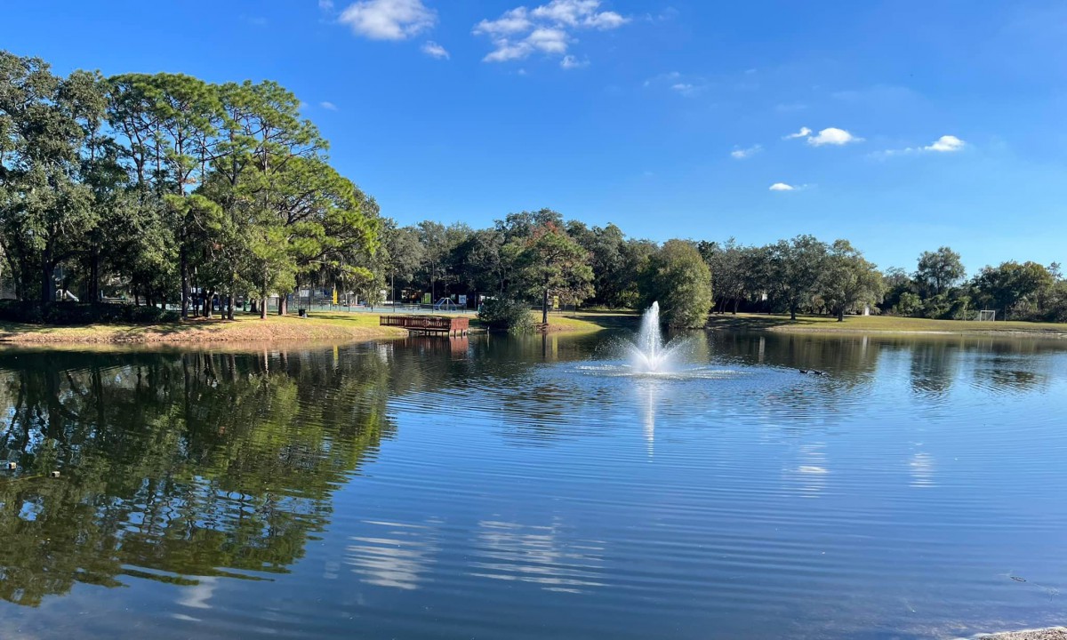 A peaceful lake with a central fountain, surrounded by lush trees under a clear blue sky.