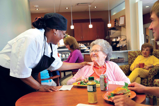 A friendly caregiver interacts with a senior diner, fostering a warm and welcoming atmosphere in a community dining setting.
