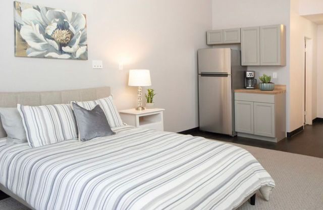 A modern bedroom featuring striped bedding, a lamp on a nightstand, and a small kitchen area with a refrigerator.