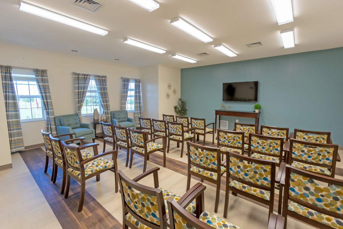 A well-lit conference room with rows of patterned chairs facing a television on the wall.