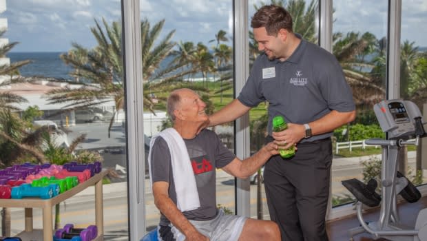 A fitness trainer supporting an elderly man during his workout session at the gym.