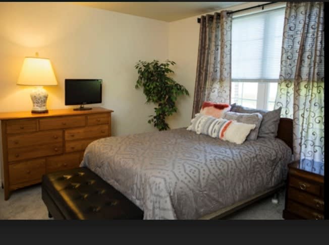 A cozy bedroom with a neatly made bed, a dresser with a TV, and soft lighting from a lamp.