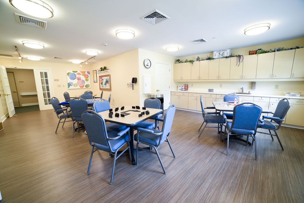 A well-lit community dining room with tables and chairs, featuring a small kitchen area.