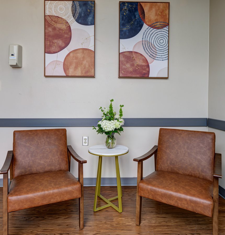 A cozy waiting area with two brown chairs, a small table adorned with flowers, and abstract artwork on the walls.