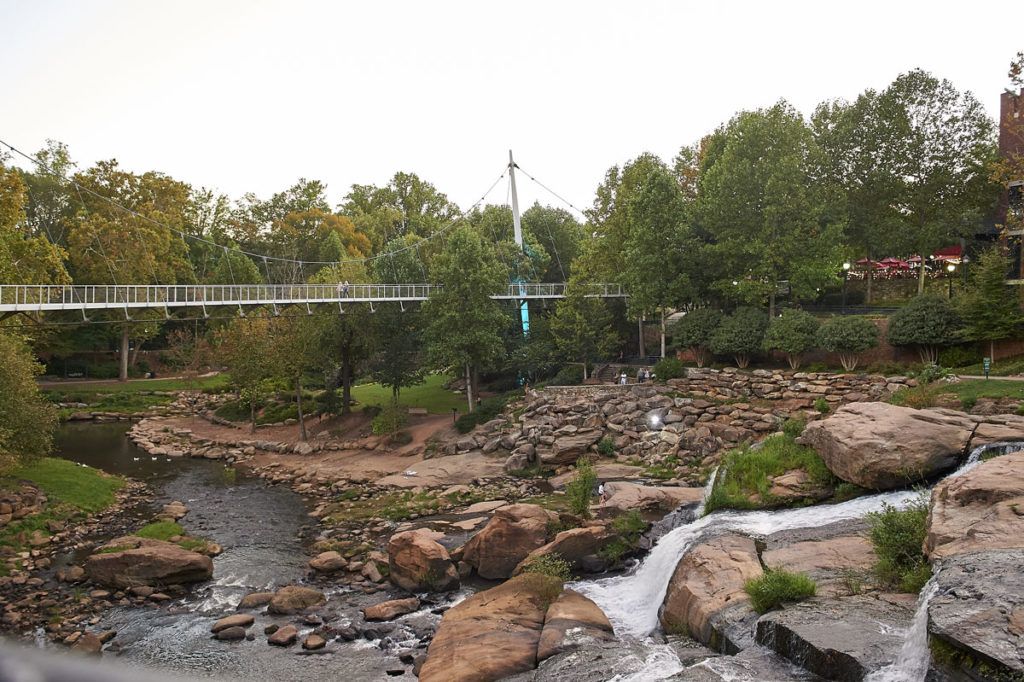A scenic view of a bridge crossing over a river with waterfalls and surrounding greenery.