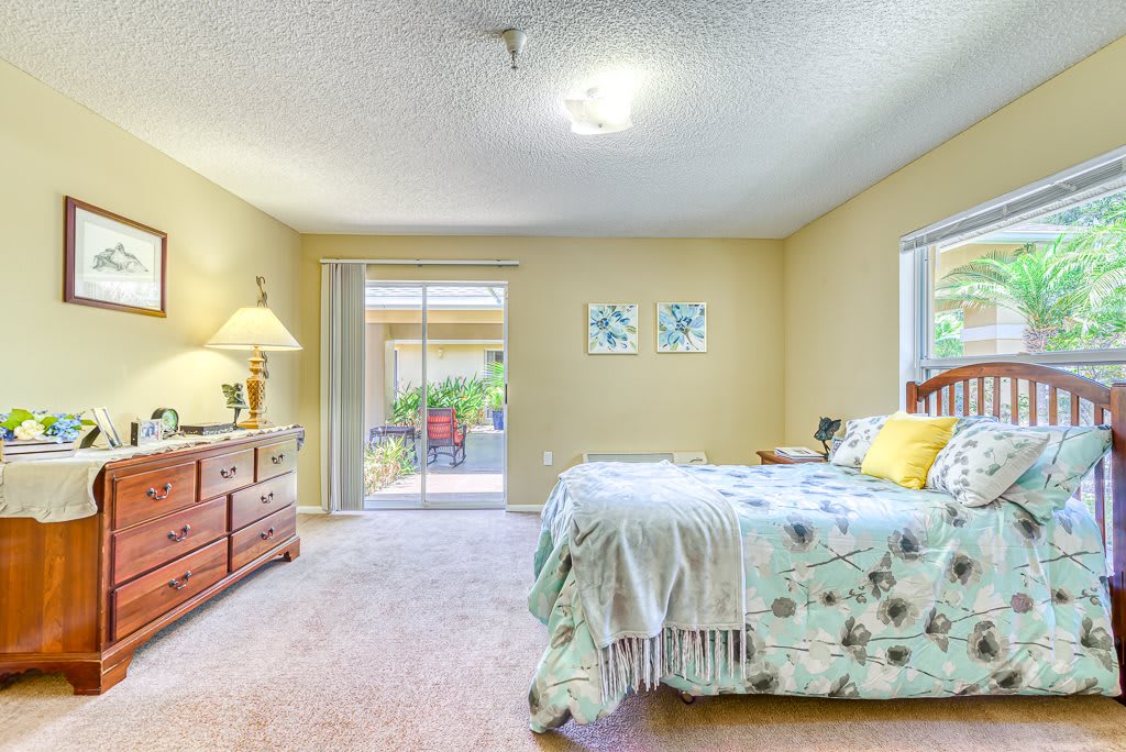 A cozy bedroom featuring a bed with floral bedding, a dresser with a lamp, and natural light filtering through the window and sliding glass door.