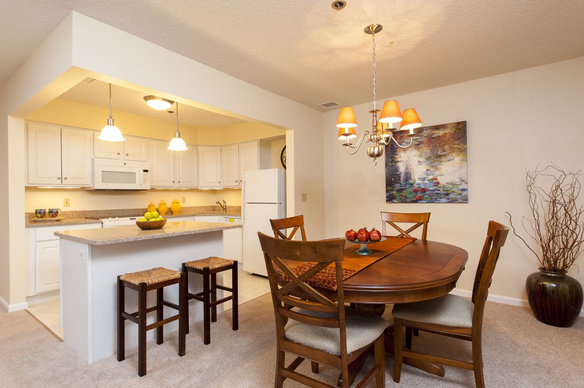 A modern dining area with a wooden table, chairs, and an open kitchen layout, featuring stylish lighting and bar stools.