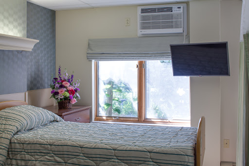 A cozy bedroom featuring a bed, vibrant flowers by the window, an air conditioning unit, and a mounted television.