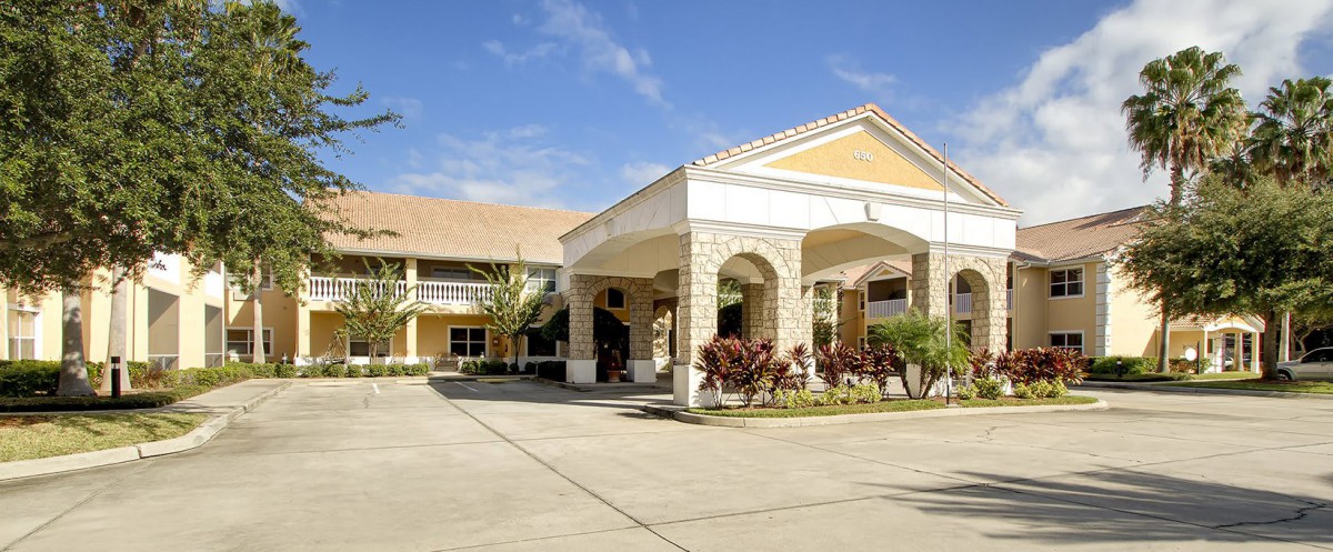 A sunny day view of a residential building with an entrance driveway, surrounded by palm trees.