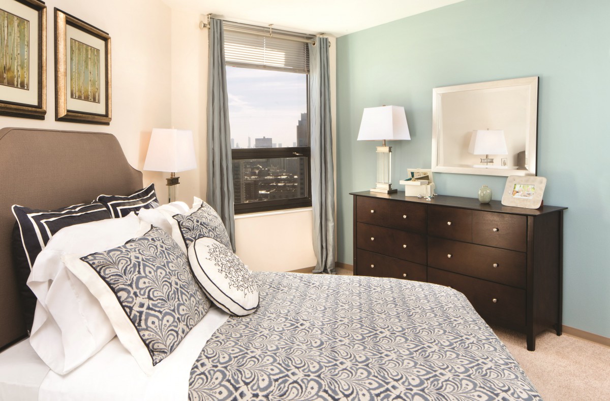 A cozy bedroom with elegant decor, featuring a bed with patterned bedding, a wooden dresser with lamps, and a window framed by curtains.