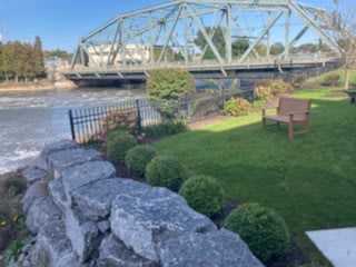 A scenic view of a bridge over a river, accompanied by a well-maintained garden with a stone wall and fencing on a sunny day.