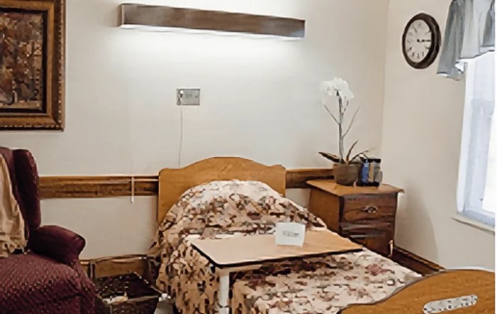 A hospital room with a single bed, floral bedding, and a nightstand with a lamp and plant.