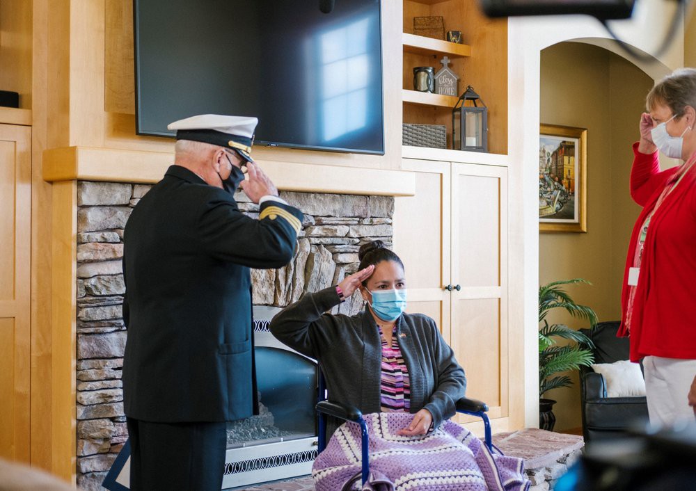 A military officer in uniform saluting a person seated in a wheelchair indoors, both wearing masks.