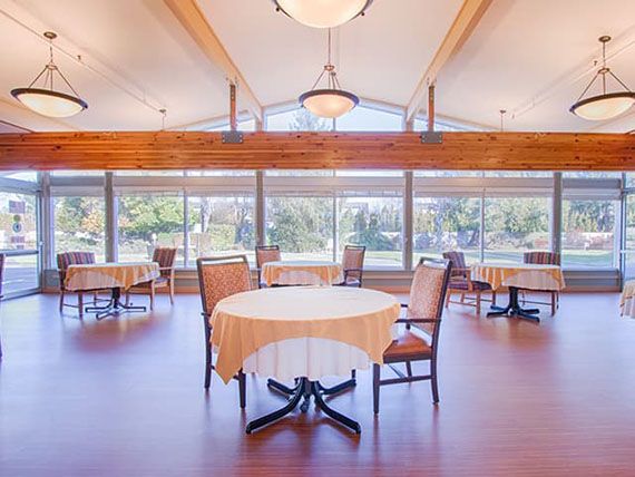 A spacious dining room with large windows and wooden flooring, featuring multiple tables and chairs.