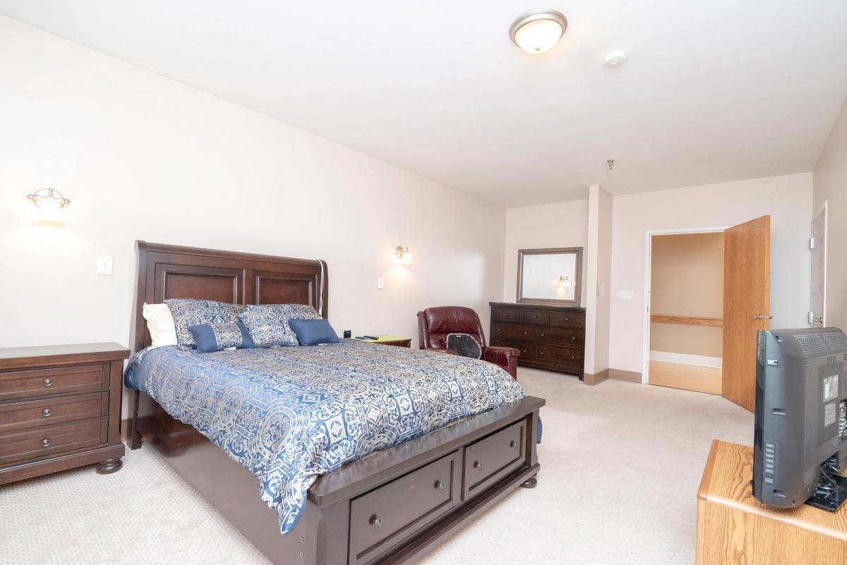 A spacious bedroom with a large bed, dresser, and chair, featuring neutral-colored walls and carpeting.