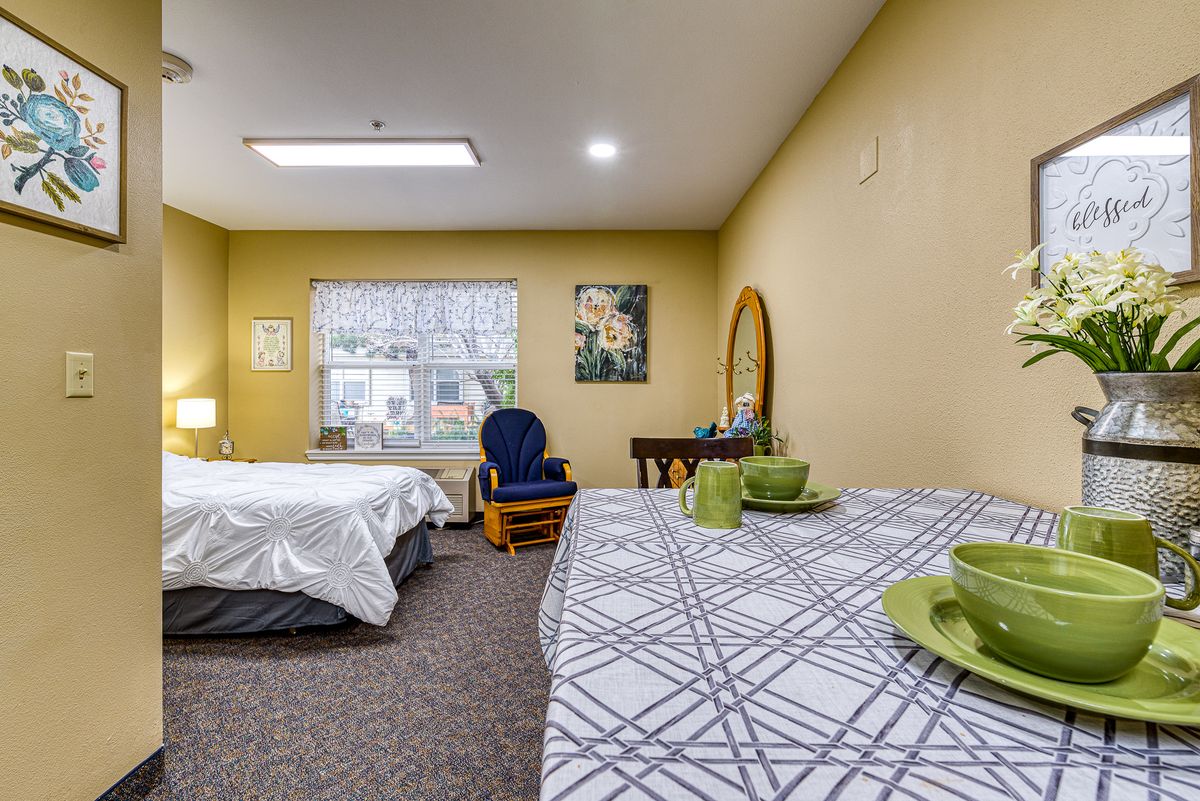 Cozy bedroom with a bed, table, and chair near a window, featuring soft lighting and simple decor.