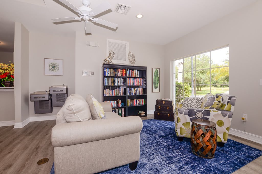A cozy living room with a sofa, armchair, and a bookshelf filled with books, featuring a large window and blue carpet.