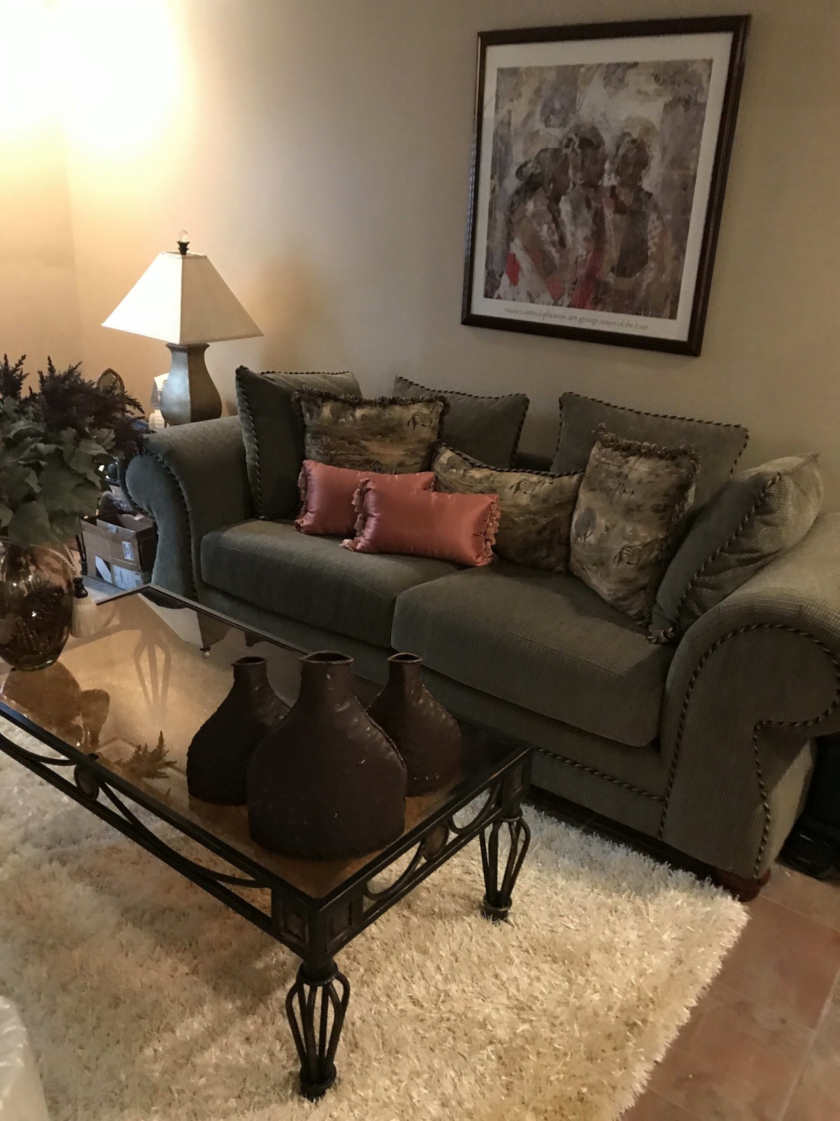 A cozy living room with a comfortable sofa, decorative cushions, and a glass coffee table adorned with vases. A lampshade adds soft lighting, and a framed painting decorates the wall.