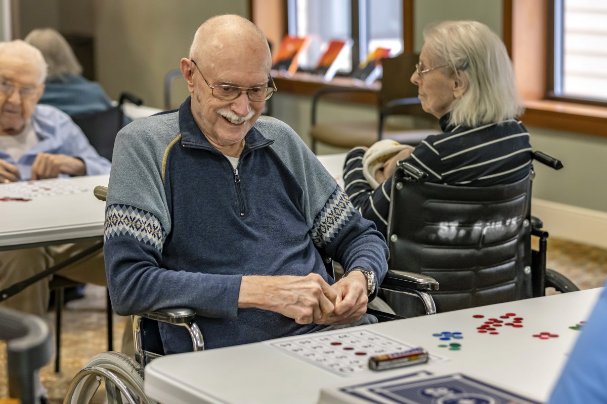 An elderly man and woman seated indoors at a table, engaging in activities and enjoying their time together.