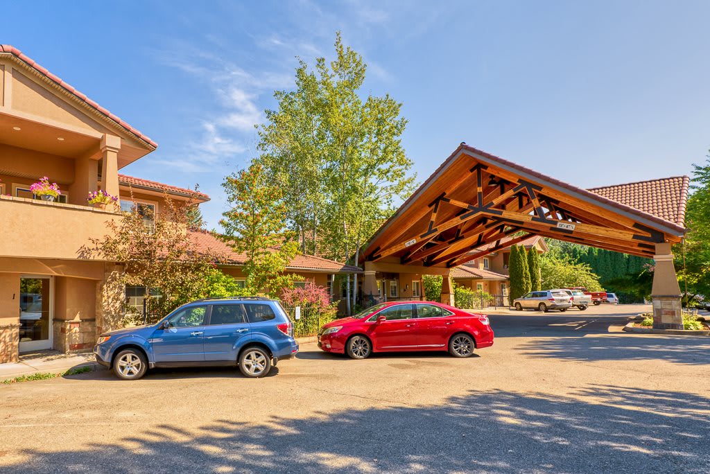 A sunny day view of a building entrance with cars parked in front, surrounded by trees.