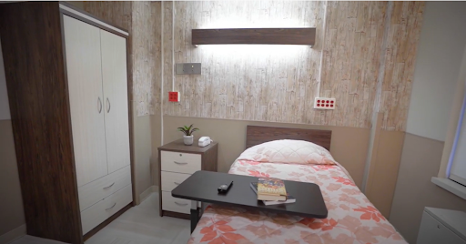 A clean and minimalist hospital room featuring a single bed, nightstand, wardrobe, and a small table.