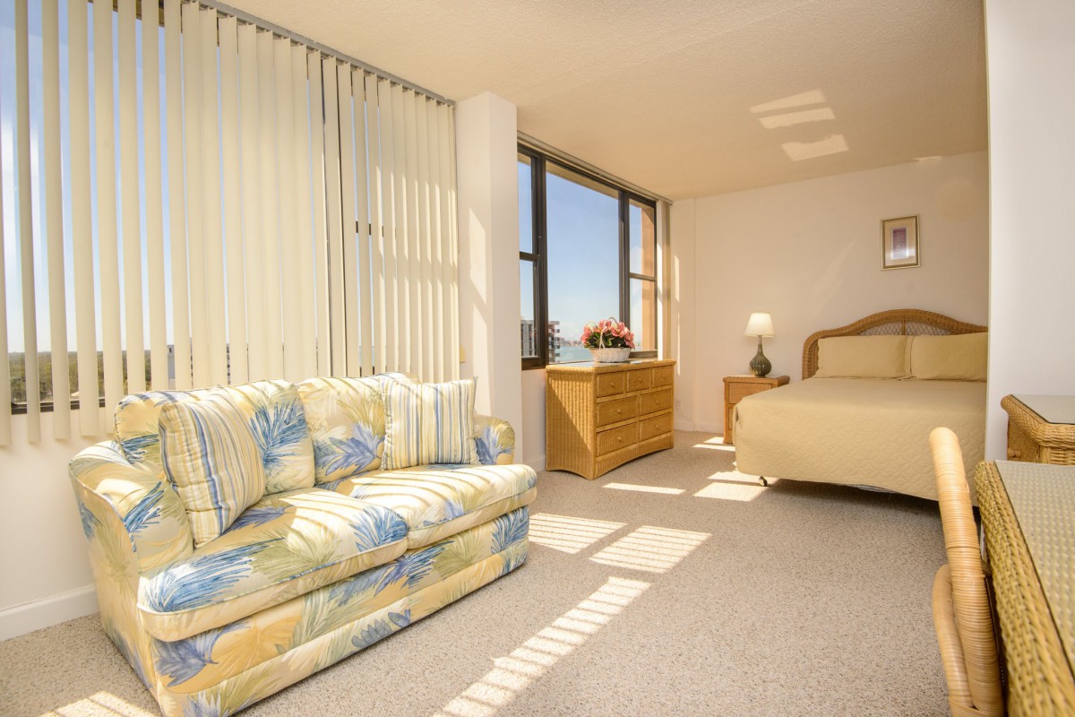 A bright bedroom with a cozy sofa, bed, and large windows letting in sunlight.
