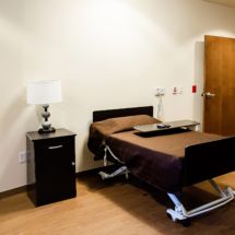 A minimalist room featuring a hospital bed, wooden flooring, and a nightstand with a lamp.