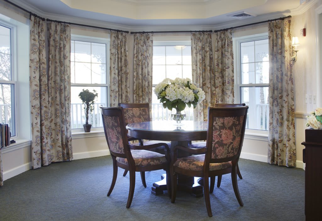 A cozy dining room with a round table, four chairs, and decorative floral arrangements beside bay windows.