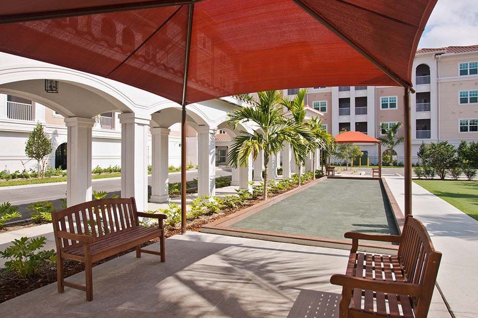 A serene outdoor patio featuring benches, a bocce court, and a red canopy with palm trees lining the walkway.