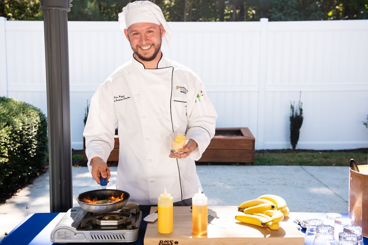 A chef in a white uniform and hat cooks outdoors, holding a bottle and a pan, with bananas on the table.