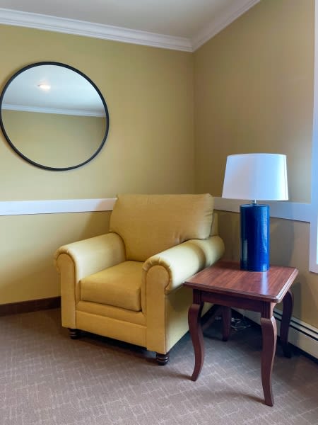 A cozy living room corner featuring a yellow armchair next to a wooden side table with a lamp, complemented by a round mirror on the wall.