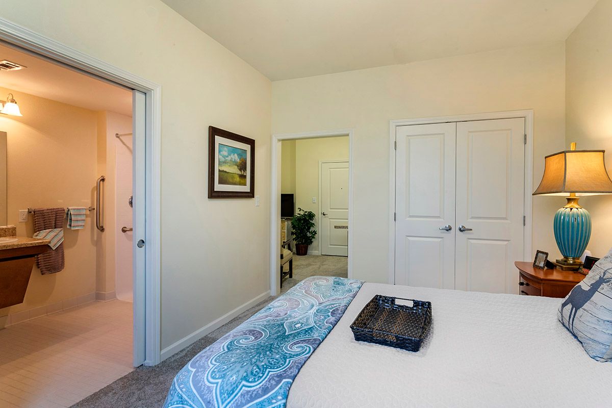 A cozy bedroom featuring a neatly made bed with a blue patterned blanket, an artwork hanging on the wall, and a view of the adjoining bathroom and closet.