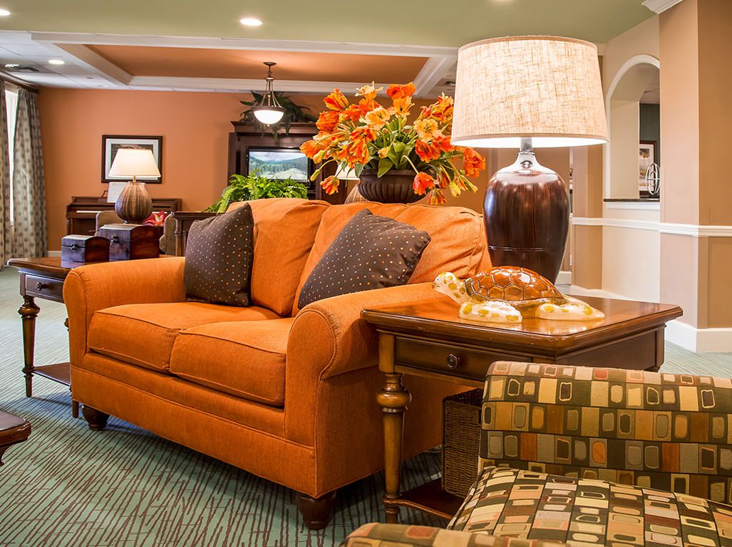A cozy living room with an orange sofa, a side table with a lamp, and a floral arrangement.