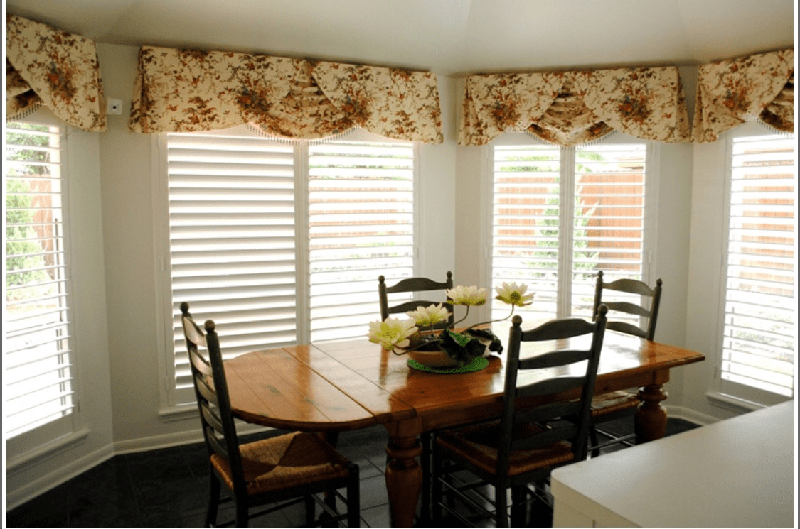 A cozy dining room with a wooden table and chairs, featuring floral curtains over large windows.