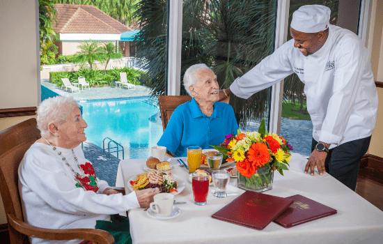A chef interacting with two seniors enjoying a meal by the pool.