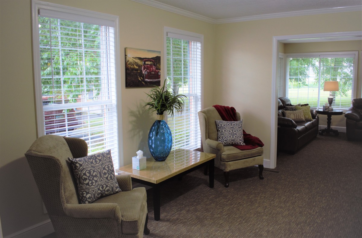 A cozy living room interior with soft chairs, a table with a decorative vase, and large windows.